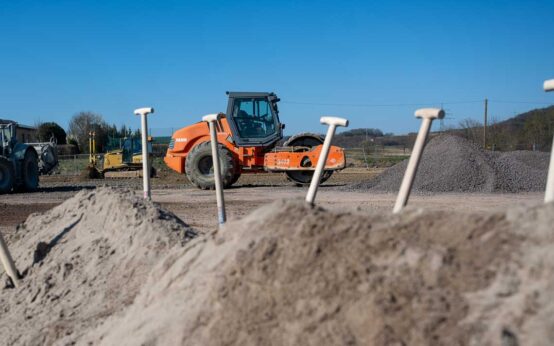 Spatenstich für neue HSK-Wohnungen, vier Spaten stecken in einem Sandhaufen, im Hintergrund ist ein Baufahrzeug.